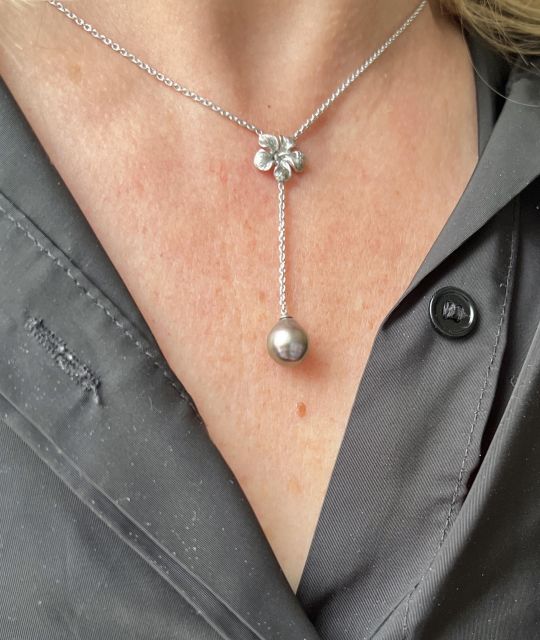 Am Kreuzungspunkt des Halses erblüht eine Blume. Eine fein geschnitzte Hibiskusblüte, die wie eine Gabe platziert ist, lässt das Licht zu einer einzigartigen Tahiti-Perle gleiten.