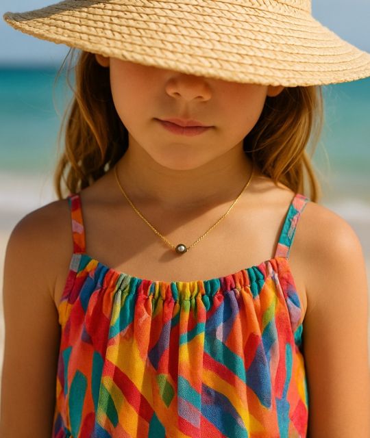 Eine Tahiti-Perle, rund und leuchtend, gleitet auf einem feinen goldenen Faden wie ein Sonnenstrahl. Eine Perlenkette für Kinder, zart wie ein Versprechen, kostbar wie die Kindheit.